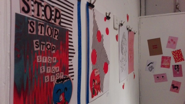 More prints on the wall and more paper hearts. on the far wall are pink postcards with cute slogans like I pug U with drawings of pug dogs. Closer are a4 prints featuring bold black lines and a vexed goofy the dog.