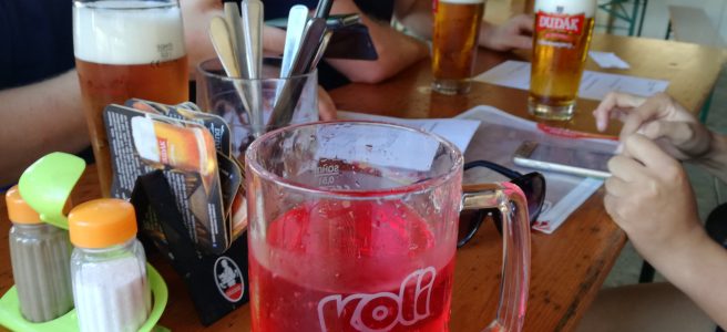 large glass of malinovka at a pub table. pints of light beer with large heads are on the table next to forks awiting food